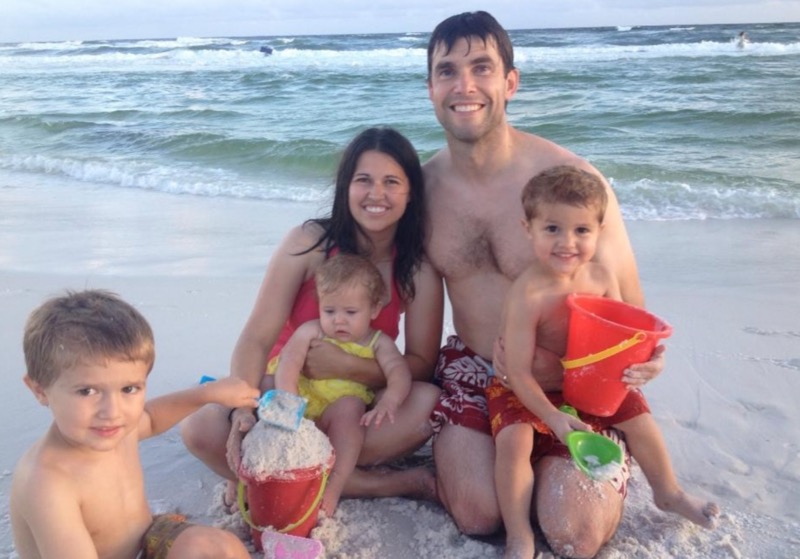 mother and father smile with two toddler boys and one toddler girl on the beach with shovels and pails