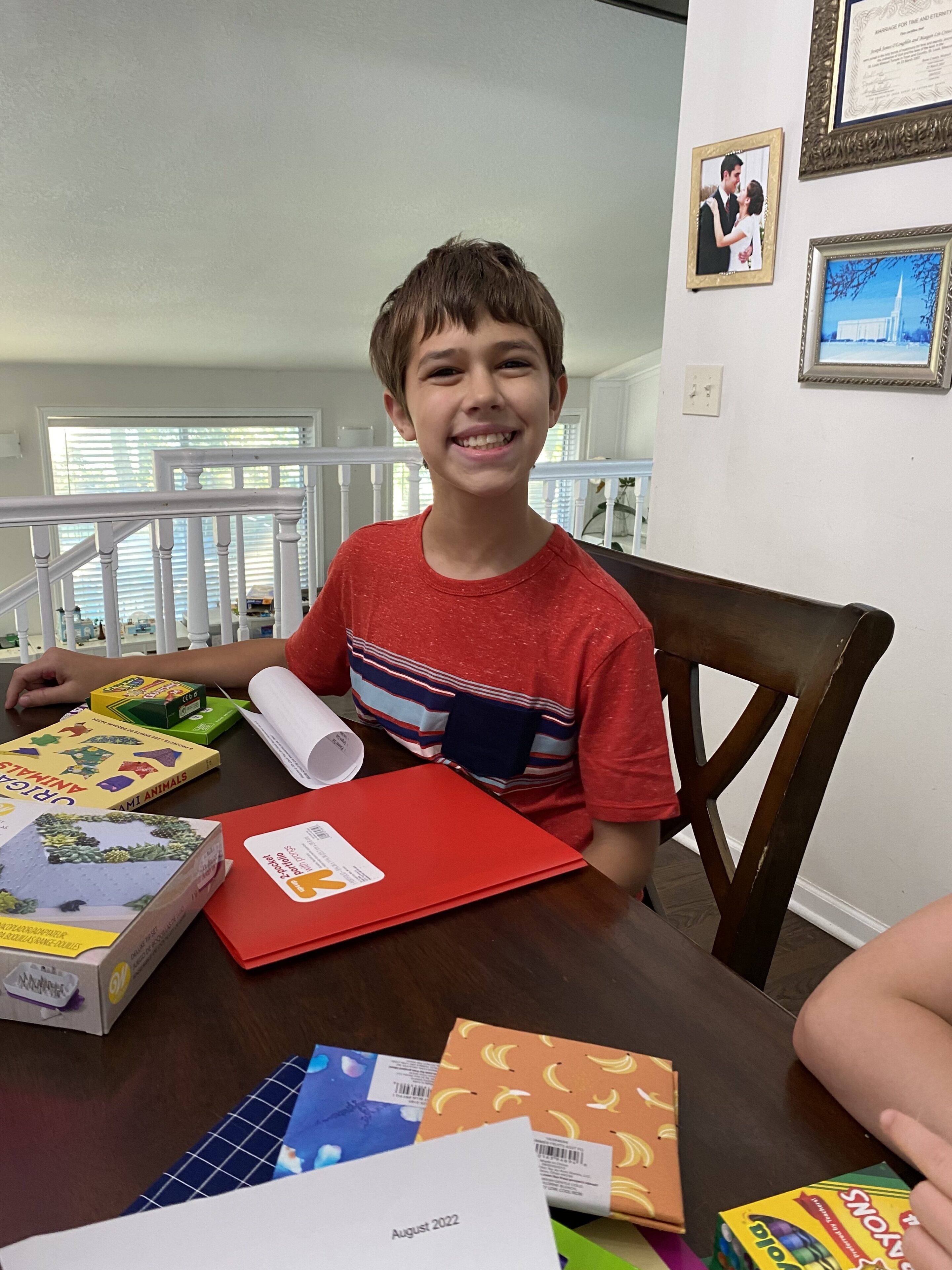 smiling boy sits at homeschool table with new supplies for first day of school tradition