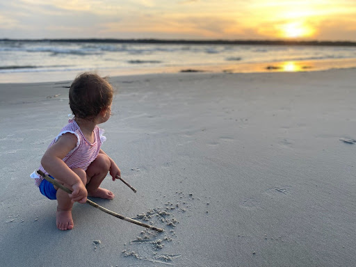 toddler draws in the sand with a stick as she looks out at the beach view